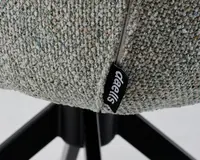 Nærbillede af en tekstureret, lysegrå polstring med flerfarvede nister, synlig syning og et sort stofmærke med hvid 'Oeells'-branding. Polstringen er monteret på en sort metalbase med skråtstillede ben.