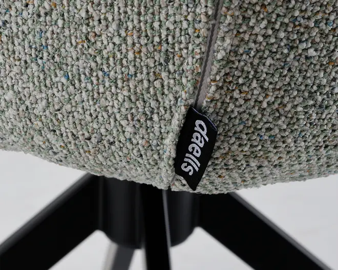 Nærbillede af en tekstureret, lysegrå polstring med flerfarvede nister, synlig syning og et sort stofmærke med hvid 'Oeells'-branding. Polstringen er monteret på en sort metalbase med skråtstillede ben.