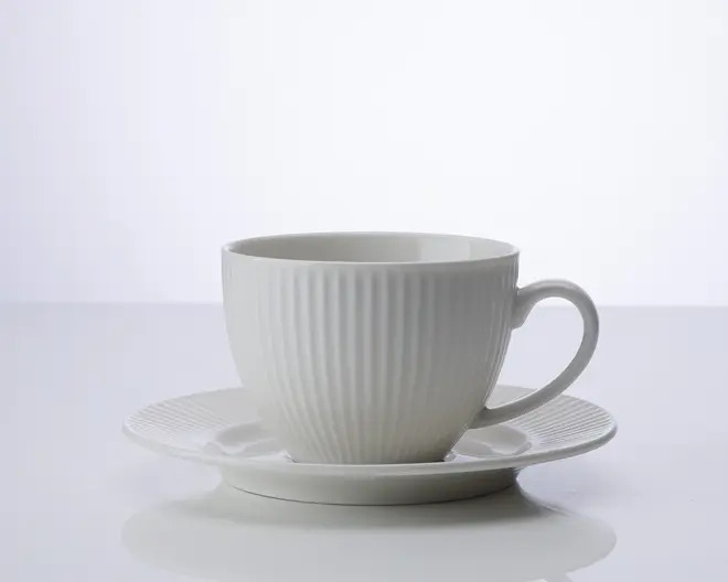 En hvid keramikkop med tilhørende underkop, begge med lodrette riller i overfladen. Koppen har et rundt håndtag og en glat, blank finish, hvilket giver et moderne og elegant udtryk.