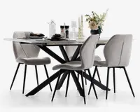Moderne spisebordssæt med et rektangulært bord med hvid marmoreffekt bordplade og sorte, vinklede metalben. Sættet inkluderer fire polstrede stole i lysebeige stof med buede rygge og slanke sorte metalben. Bordet er pyntet med glas, tallerkener, lys og en blomsterdekoration i en mørk vase, hvilket giver et elegant og nutidigt udtryk.