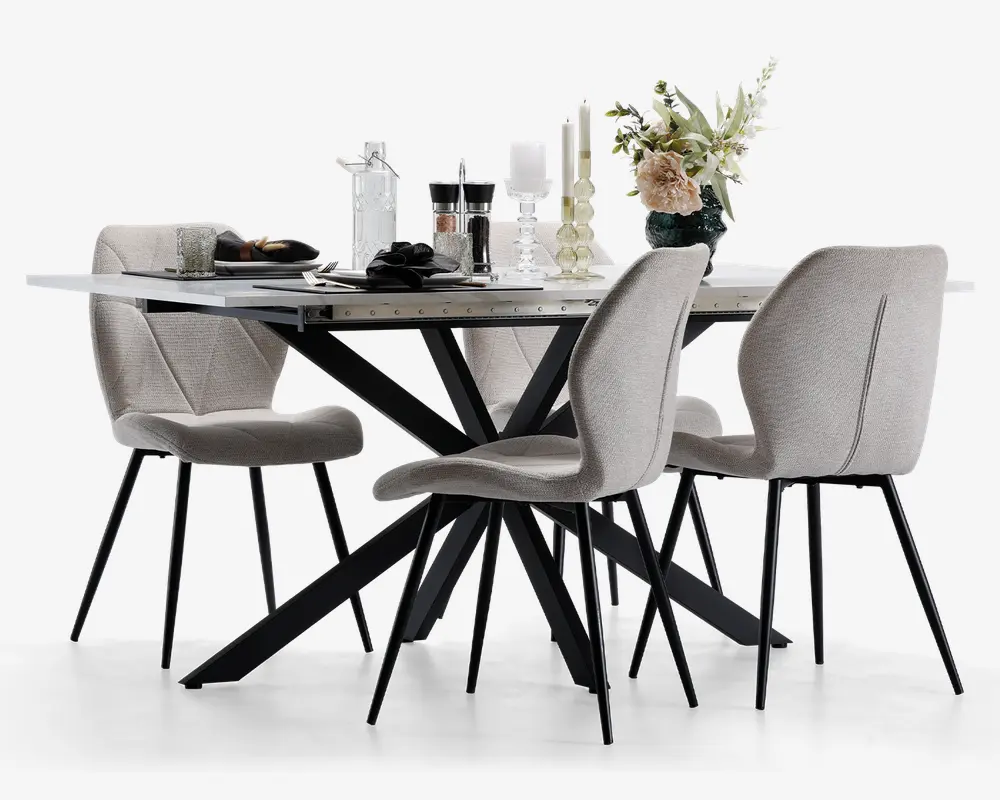 Moderne spisebordssæt med et rektangulært bord med hvid marmoreffekt bordplade og sorte, vinklede metalben. Sættet inkluderer fire polstrede stole i lysebeige stof med buede rygge og slanke sorte metalben. Bordet er pyntet med glas, tallerkener, lys og en blomsterdekoration i en mørk vase, hvilket giver et elegant og nutidigt udtryk.