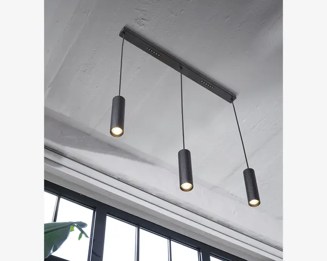 Loftmonteret lampe med tre cylindriske pendler, der hænger fra en rektangulær metalbase. Hver pendel har en rillet sort overflade og udsender et varmt lys nedad. Ledningerne er jævnt fordelt, hvilket giver et moderne og minimalistisk udtryk, der passer til nutidige indretninger.