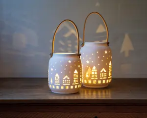 To hvide lanterner af keramik med udskårne motiver af huse, træer og stjerner. Lanternerne udsender et varmt, gult lys indefra, som oplyser de udskårne mønstre og kaster dekorative skygger på væggen. Hver lanterne har et buet metalhåndtag, så de er nemme at bære eller hænge op. Overfladen er glat og mat, og lanternerne står på en træoverflade.