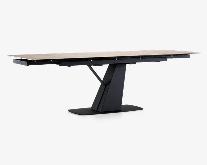 Rektangulært bord med en lys, glat bordplade og en moderne, vinklet sort sokkel. Soklen har et geometrisk design med en bred, flad bund for stabilitet og en central støtte, der vinkler opad for at møde bordpladen. Bordet ser ud til at have en udtræksmekanisme, som giver mulighed for ekstra siddepladser eller større overflade.