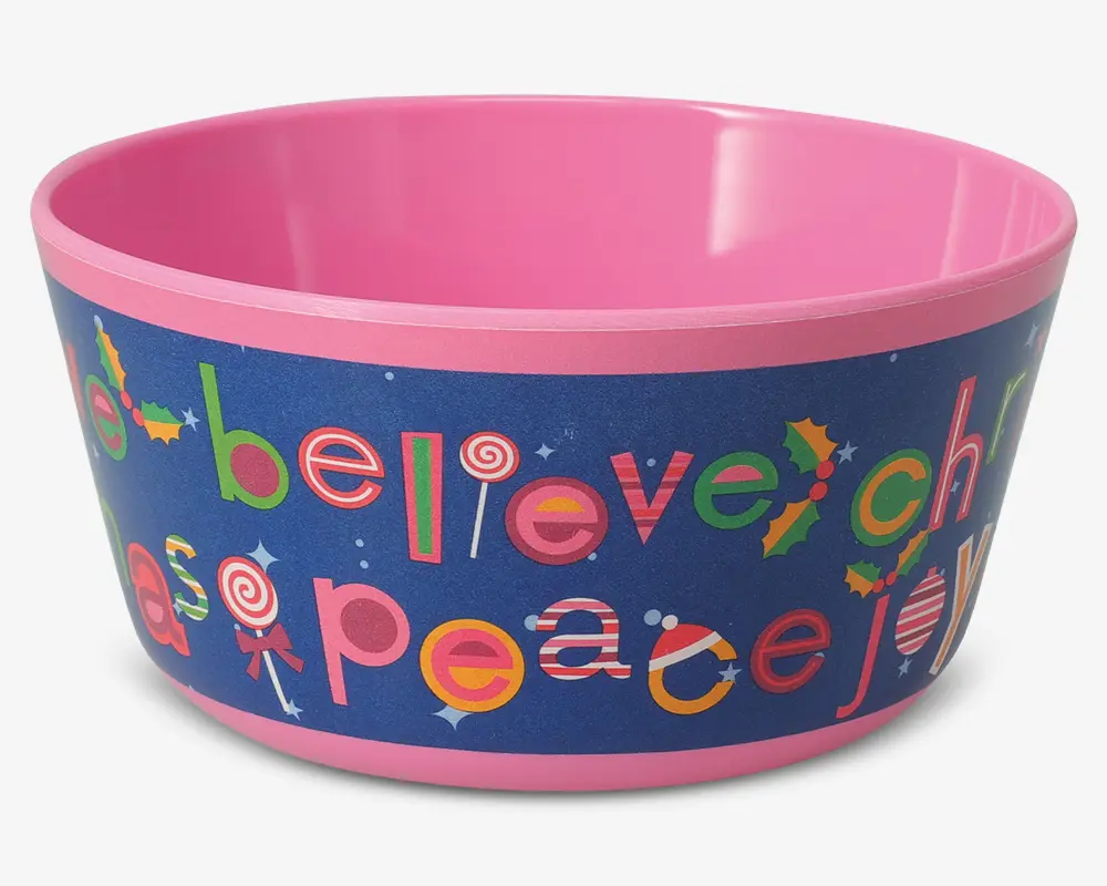 En rund, lyserød skål med en blå ydre kant, der har farverige, festlige bogstaver og juleinspirerede motiver. Ordene 'believe', 'peace' og 'joy' er skrevet med legende skrifttyper, hvor nogle bogstaver er dekoreret som slikstokke, slikkepinde, kristtornblade og stjerner. Skålen har en glat, blank overflade og en bred åbning, velegnet til servering af snacks eller godter.