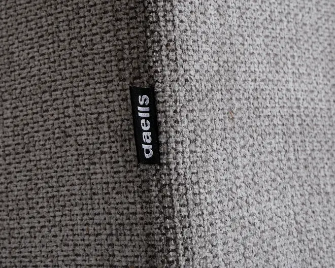 Nærbillede af et lysegråt, vævet tekstil med en tydelig lodret syning. En lille sort stofmærkat med mærket 'daells' i hvide bogstaver er syet ind i syningen. Stoffet ser slidstærkt ud og har et diskret, ternet mønster.