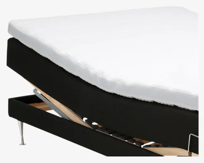Nærbillede af en seng med en hvid madras og sort polstret bund. Sengerammen er delvist løftet, så man kan se et opbevaringsrum under sengen. Rammen har metalben og lameller af træ til at støtte madrassen.