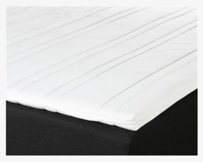 Nærbillede af en hvid topmadras med glat overflade og diskrete, jævnt fordelte, diagonale syninger. Topmadrassen ser tynd og blød ud og har en kant, der ligger pænt oven på en sort madrasbund.