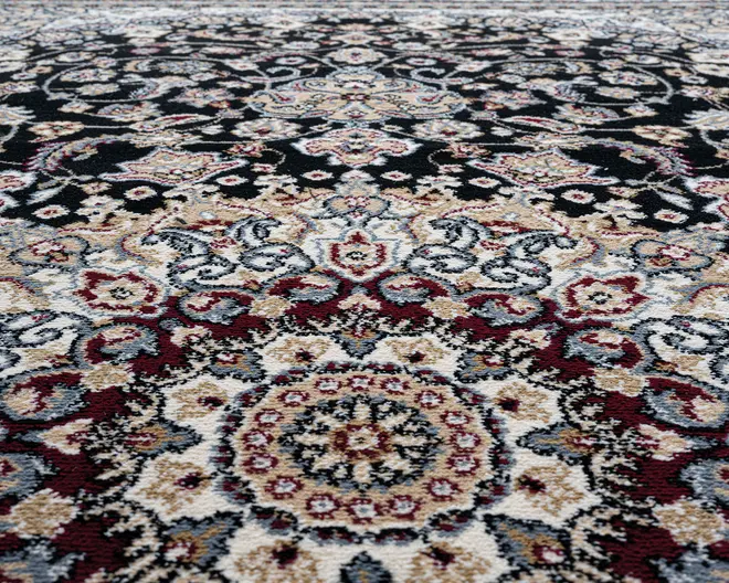 Nærbillede af et tæppe med et indviklet blomstermønster og medaljon-design. Mønsteret består af detaljerede motiver i bordeaux, creme, sort, grå og beige nuancer. Teksturen ser blød og tæt ud, hvilket fremhæver de dekorative detaljer og farvekontraster.