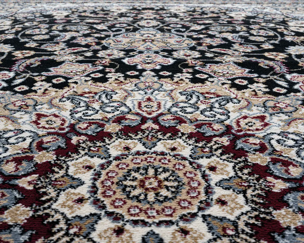 Nærbillede af et tæppe med et indviklet blomstermønster og medaljon-design. Mønsteret består af detaljerede motiver i bordeaux, creme, sort, grå og beige nuancer. Teksturen ser blød og tæt ud, hvilket fremhæver de dekorative detaljer og farvekontraster.