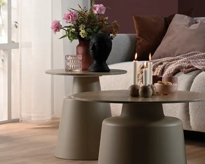 To runde pedestalborde i en mat taupe-finish, det ene højere og mindre og det andet bredere og lavere, stylet ved siden af en sofa med lys, et glas, blomster og dekorative genstande.