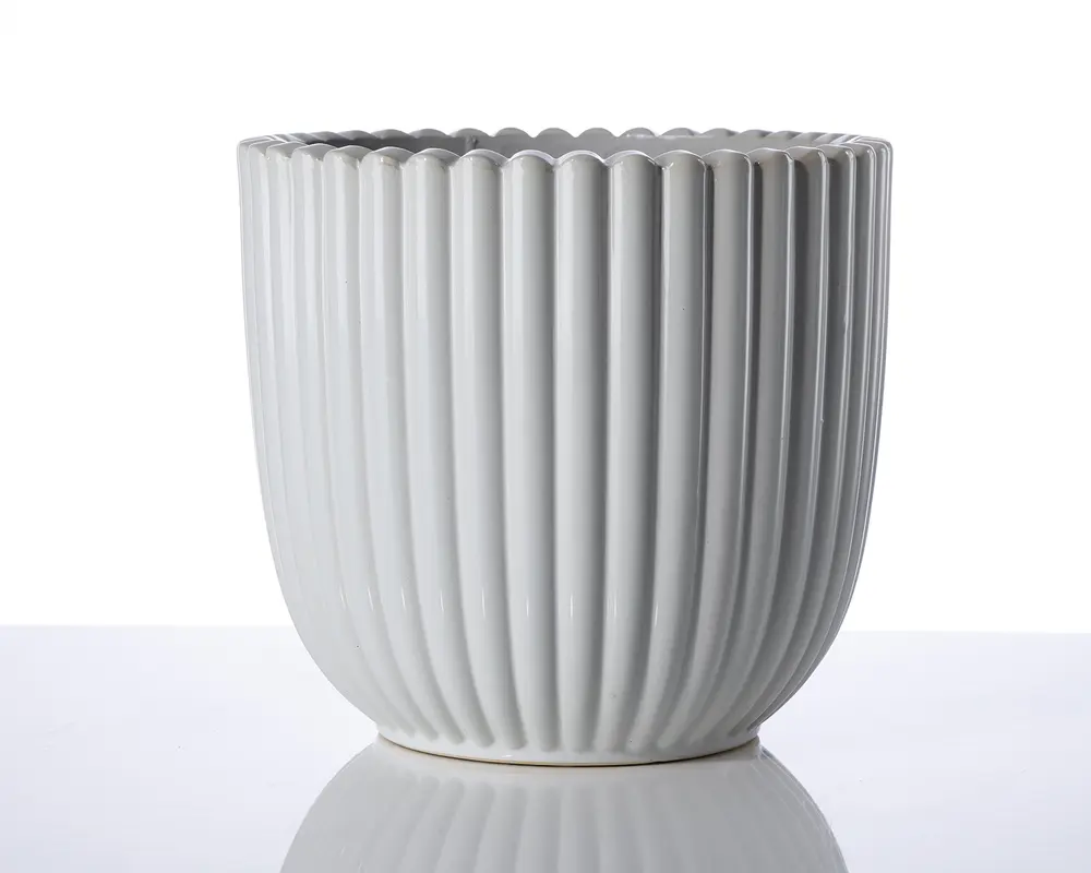 En hvid keramisk beholder med lodrette riller og bølget kant. Overfladen er blank, og formen er cylindrisk med en let afrundet bund. Designet er moderne og minimalistisk, velegnet til dekorative formål.