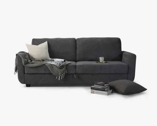 En moderne sofa i mørkegråt stof med rene linjer og brede armlæn. Sofaen har to store sædehynder og to ryghynder for komfort. På venstre side ligger en cremefarvet pyntepude og et sort-hvidt mønstret tæppe over armlænet. Et åbent magasin ligger på sædet. På højre side står en lille kop og underkop på sædehynden. Foran sofaen er der en stak bøger og en stor mørkegrå gulvpude.