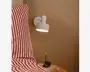 En moderne vægmonteret lampe med mat hvid finish, cylindrisk lampeskærm og en lille cylindrisk detalje på toppen. Lampen er placeret over et sengebord ved siden af en seng med rød- og hvidstribet sengetøj. Lampen giver et fokuseret nedadrettet lys, ideelt til læsning eller brug ved sengen.