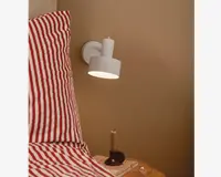 En moderne vægmonteret lampe med mat hvid finish, cylindrisk lampeskærm og en lille cylindrisk detalje på toppen. Lampen er placeret over et sengebord ved siden af en seng med rød- og hvidstribet sengetøj. Lampen giver et fokuseret nedadrettet lys, ideelt til læsning eller brug ved sengen.
