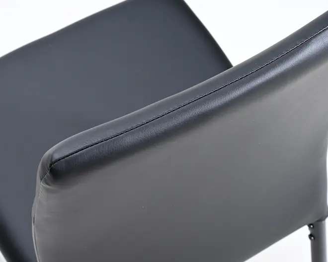 Nærbillede af en stol med glat, mat sort kunstlæderpolstring. Ryglænet er let buet for ergonomisk støtte og har synlige, pæne syninger langs den øverste kant. Sædet ser også polstret ud for ekstra komfort, og designet er moderne og minimalistisk.