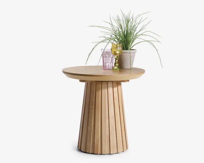 Rundt sidebord med naturtræ-finish og en rillet, konisk sokkel. Bordpladen er glat og cirkulær. Ovenpå står en dekorativ opsætning bestående af en potteplante med lange, tynde grønne blade, et lyserødt facetslebet glas og et guldfarvet vinglas, hvilket giver et elegant og stilfuldt udtryk.