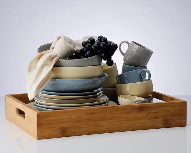 Et sæt keramikservice i afdæmpede nuancer af blå, grå og beige, herunder tallerkener, skåle og krus, pænt stablet i en rektangulær bakke af træ. Sættet har en blanding af blanke og matte overflader. En cremefarvet stofserviet er lagt over den øverste skål, og en klase kunstige sorte druer er placeret øverst som dekoration.