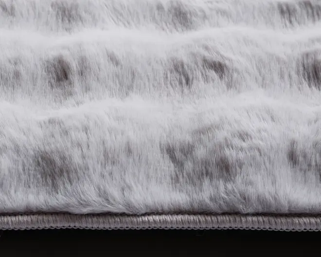 Nærbillede af et blødt, plysset stof med en imiteret pelsstruktur. Materialet har et bølget, rillet mønster og en blanding af lysegrå og mørkere grå nuancer, hvilket giver et naturligt og luksuriøst udseende. Kanten er pænt afsluttet med en syet kant for ekstra holdbarhed.