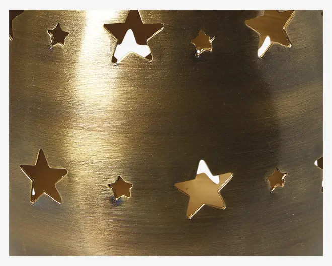Nærbillede af en metaloverflade med børstet guldfinish, der har flere udskæringer i form af stjerner i forskellige størrelser. Overfladen er blank og reflekterende, og udskæringerne lader lys passere igennem for et dekorativt udtryk.