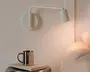 Minimalistisk hvid vægmonteret justerbar lampe med rund base, vinklet arm og lille cylindrisk skærm, vist tændt ved siden af en seng og et natbord.