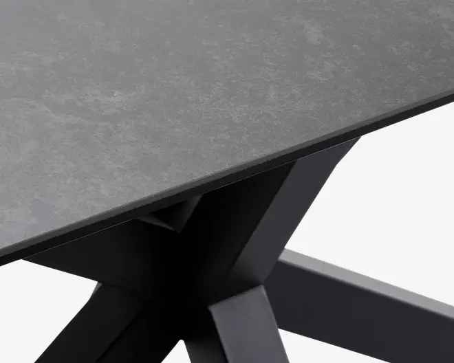 Nærbillede af et bordhjørne med en mat, stenlignende mørkegrå bordplade med tynd profil. Bordet har en moderne, vinklet sort metalbase med krydsende ben, hvilket giver et robust og nutidigt udtryk.