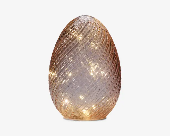 Dekorativt lys formet som et æg med en tekstureret glasoverflade i spiral-mønster. Glasset har en let ravfarvet tone og indeholder små, varme hvide LED-lys indeni, som giver en blød, glødende effekt. Basen er flad, så den står stabilt på overflader.