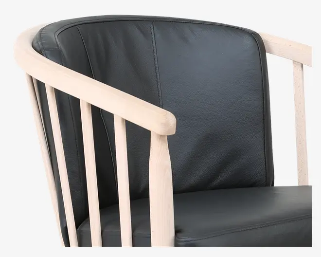 Nærbillede af en stol med glat, sort lædersæde og ryghynde. Stellet er lavet af lyst træ med en naturlig finish, inklusive afrundede armlæn og lodrette spindler. Designet kombinerer moderne og klassiske elementer og giver både komfort og stil.
