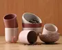 Et sæt keramiske kopper og skåle i afdæmpede rosa, beige og marmorerede nuancer. Produkterne har en mat overflade og et minimalistisk design, hvor nogle kopper har tofarvet udseende. En kop har en tepose med mærkat, der hænger ud over kanten, og en skål indeholder kaffebønner, mens flere bønner ligger spredt på træbordet. Formerne er enkle og afrundede, velegnet til servering af varme drikke eller snacks.