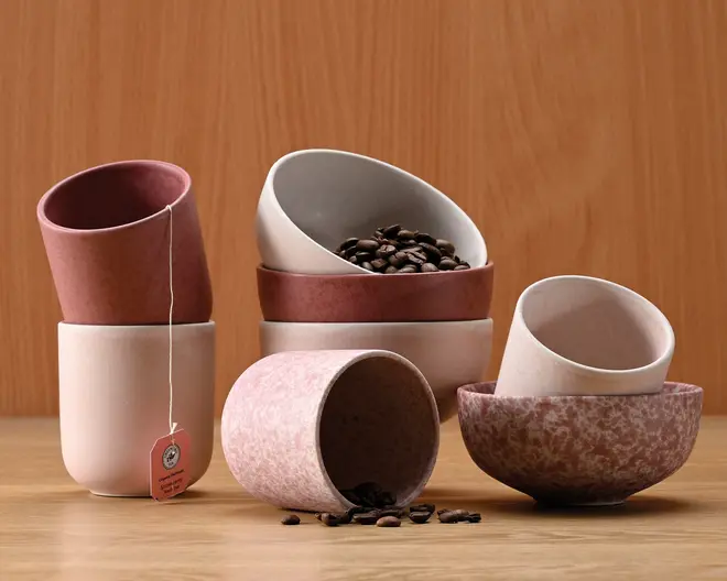 Et sæt keramiske kopper og skåle i afdæmpede rosa, beige og marmorerede nuancer. Produkterne har en mat overflade og et minimalistisk design, hvor nogle kopper har tofarvet udseende. En kop har en tepose med mærkat, der hænger ud over kanten, og en skål indeholder kaffebønner, mens flere bønner ligger spredt på træbordet. Formerne er enkle og afrundede, velegnet til servering af varme drikke eller snacks.