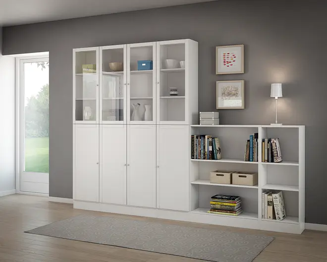 Høj, hvid opbevaringsenhed med en kombination af overskabe med glaslåger og underskabe med solide låger, samt åbne hylder. Glaslågerne viser pænt arrangerede dekorationsgenstande og bokse, mens de åbne hylder indeholder bøger, opbevaringskurve og ekstra dekoration. Enheden har rene linjer og et moderne design, velegnet til organisering og udstilling af ting i stuen.