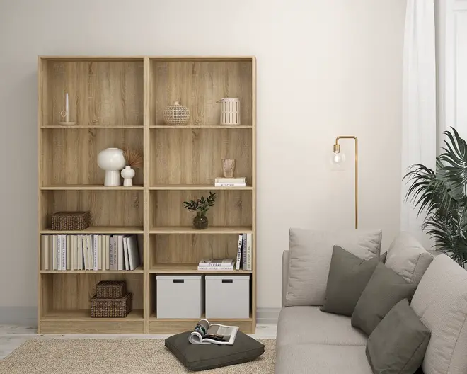 Høj reol i lyst træ med fem åbne hylder. Hylderne er dekoreret med forskellige pyntegenstande, herunder en hvid moderne lampe, flettede kurve, keramikvaser, bøger placeret både lodret og vandret samt to hvide opbevaringskasser nederst til højre. Reolen har en naturlig træstruktur og et minimalistisk, skandinavisk-inspireret design.