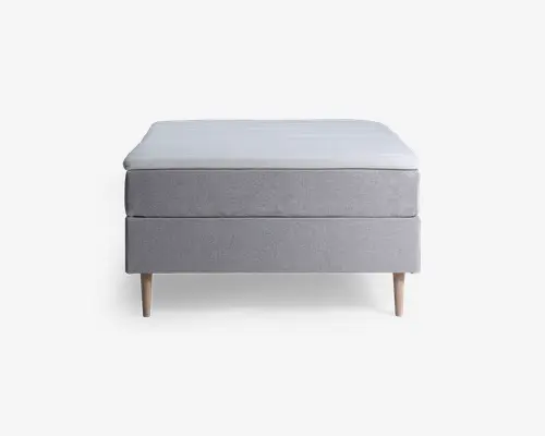 Minimalistisk seng med en lysegrå polstret bund og madras, understøttet af fire lyse træben. Designet er enkelt og moderne med rene linjer og en lav profil. Stoffet fremstår glat og stramt monteret, hvilket giver et pænt og nutidigt udtryk.