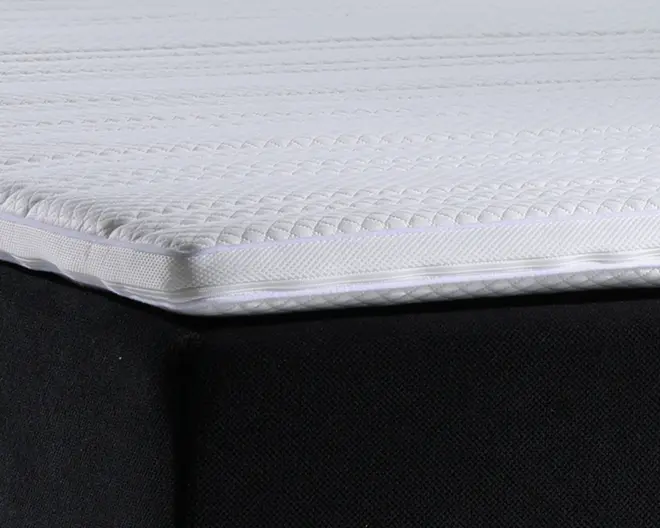 Nærbillede af et madras-hjørne, der viser en hvid, quiltet og tekstureret overflade med et tykt, polstret lag. Syningen er synlig langs kanten, og madrassen ligger oven på en sort stofbetrukket bund. Overfladen ser blød og polstret ud, hvilket indikerer komfort og støtte.