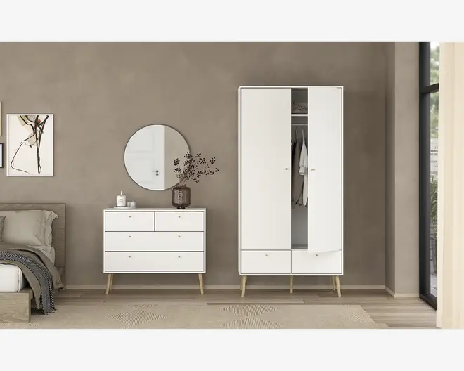 Billedet viser et moderne soveværelsesmiljø med en hvid kommode med fire skuffer og skråtstillede træben samt et matchende hvidt garderobeskab med to låger, to nederste skuffer og tilsvarende træben. Garderobeskabet har den ene låge let åben, så man kan se hængende tøj indeni. På kommoden står dekorative genstande, herunder et rundt spejl, en vase med grene og små beholdere. Møblerne har et minimalistisk design med rene linjer og små runde greb, placeret mod en neutral taupefarvet væg og lyst trægulv.