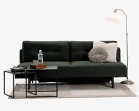 En moderne mørkegrøn sofa med rene linjer og minimalistisk design, udstyret med to bløde ryghynder. Sofaen har en rund, plisseret grå pude og en stor, fluffy cremefarvet pude for ekstra komfort. Den står på slanke sorte metalben. Foran sofaen er der to indskudsborde i sort metal; det ene har en glaskaraffel, to glas og en træbakke, mens det andet har en stak bøger. Opstillingen fuldendes af et neutralt gulvtæppe og en høj gulvlampe i messing med en hvid plisseret lampeskærm.
