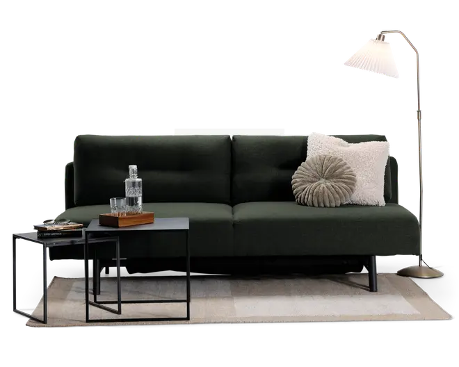 En moderne mørkegrøn sofa med rene linjer og minimalistisk design, udstyret med to bløde ryghynder. Sofaen har en rund, plisseret grå pude og en stor, fluffy cremefarvet pude for ekstra komfort. Den står på slanke sorte metalben. Foran sofaen er der to indskudsborde i sort metal; det ene har en glaskaraffel, to glas og en træbakke, mens det andet har en stak bøger. Opstillingen fuldendes af et neutralt gulvtæppe og en høj gulvlampe i messing med en hvid plisseret lampeskærm.