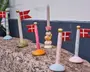 En samling dekorative flagholdere, hver med forskelligt farvede og mønstrede baser, der bærer små Dannebrog-flag med rød baggrund og hvidt kors. Basen er rund og findes i farver som blå, grøn, orange, pink og hvid med gulddetaljer. Én holder i midten er designet til at holde et lys og er udsmykket med en fantasifuld figur af en gul fugl og en lyserød blomst. Produkterne er placeret på en stensokkel foran en blå og hvid væg.