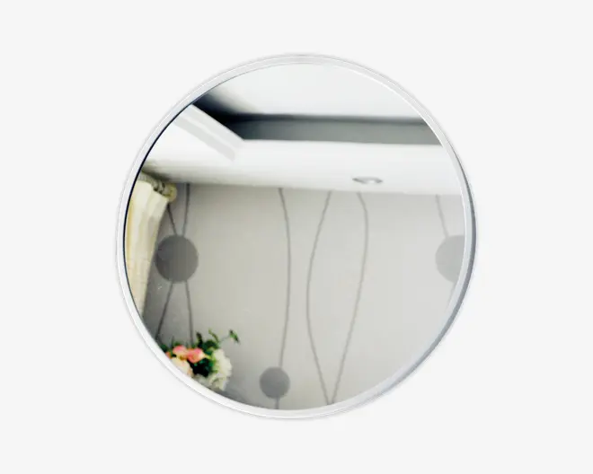 Rundt spejl med en tynd, sølvfarvet ramme. Spejlbilledet viser et moderne interiør med lyse vægge, der har et diskret, abstrakt mønster af linjer og cirkler. Til venstre ses et gardin og nederst til venstre et lille blomsterarrangement.