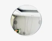 Rundt spejl med en tynd, sølvfarvet ramme. Spejlbilledet viser et moderne interiør med lyse vægge, der har et diskret, abstrakt mønster af linjer og cirkler. Til venstre ses et gardin og nederst til venstre et lille blomsterarrangement.