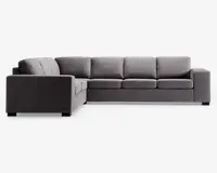 Moderne L-formet hjørnesofa med venstrevendt chaiselong, polstret i mørkegråt stof. Sofaen har brede, firkantede armlæn, tykke sædehynder og bløde ryghynder for ekstra komfort. Designet er minimalistisk med rene linjer, velegnet til moderne stuer.