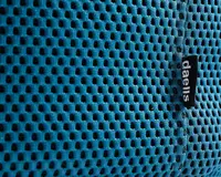 Nærbillede af et blåt mesh-stof med jævnt fordelte, små ovale huller for åndbarhed. Materialet ser blødt og fleksibelt ud, velegnet til ventilation. Et sort stofmærke med hvid tekst 'daells' er syet ind i sømmen og angiver mærket.