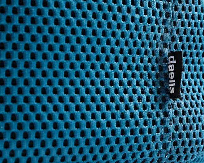 Nærbillede af et blåt mesh-stof med jævnt fordelte, små ovale huller for åndbarhed. Materialet ser blødt og fleksibelt ud, velegnet til ventilation. Et sort stofmærke med hvid tekst 'daells' er syet ind i sømmen og angiver mærket.