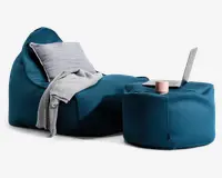 Et moderne loungemiljø med en dyb teal-farvet, tekstureret sækkestol og en matchende puf. Sækkestolen er udstyret med en lysegrå pude og et blødt, gråt tæppe, der er draperet over stolen. Puffen fungerer som et sidebord og har en lyserød kop, en lukket notesbog og en åben bærbar computer ovenpå. Stoffet har et vævet, netlignende udseende, og helhedsindtrykket er hyggeligt og indbydende.