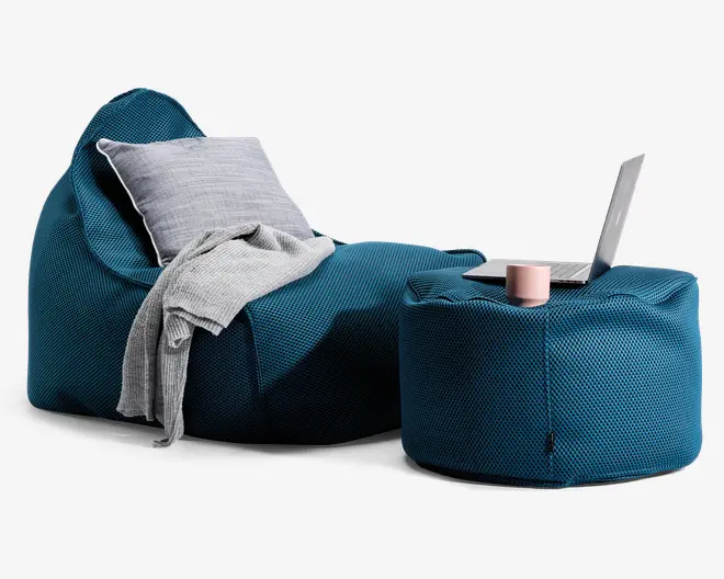 Et moderne loungemiljø med en dyb teal-farvet, tekstureret sækkestol og en matchende puf. Sækkestolen er udstyret med en lysegrå pude og et blødt, gråt tæppe, der er draperet over stolen. Puffen fungerer som et sidebord og har en lyserød kop, en lukket notesbog og en åben bærbar computer ovenpå. Stoffet har et vævet, netlignende udseende, og helhedsindtrykket er hyggeligt og indbydende.