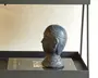 En dekorativ buste-skulptur lavet af mørkt, mat materiale, sandsynligvis keramik eller resin. Bustens detaljer omfatter et tekstureret hovedbeklædning med en hævet kant og et mønstret krave omkring halsen. Den overordnede stil antyder et vintage eller klassisk design med glatte konturer og diskret ornamentering. Skulpturen står på en sort bakke oven på en bog, hvilket giver et sofistikeret udtryk til udstillingen.
