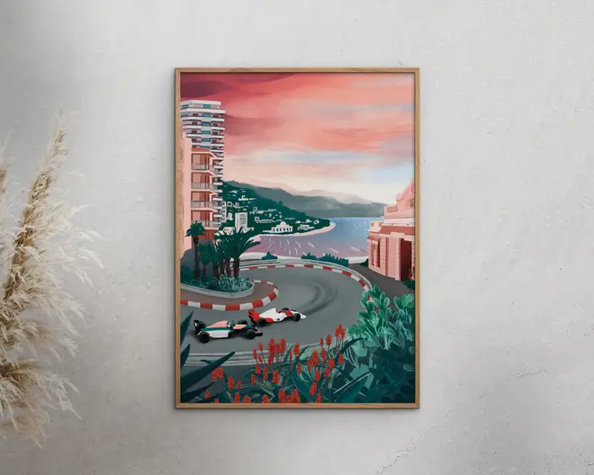 Indrammet kunstværk med en stiliseret illustration af et kystbylandskab ved solnedgang, hvor to Formel 1-racerbiler kører gennem et skarpt sving på en gadebane. Scenen viser moderne højhuse, frodig beplantning med røde blomster i forgrunden, palmer samt udsigt over havet og fjerne bakker under en lyserød og orange himmel.