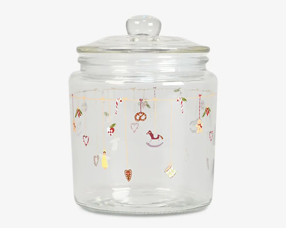 Klar glasbeholder med rundt låg og knop som håndtag. Beholderen er dekoreret med små, farverige illustrationer med juletema, såsom slikstokke, hjerter, kristtorn, gyngehest, pretzel, engel og andre festlige motiver, der hænger fra tynde gyldne linjer rundt om den øverste del af glasset.