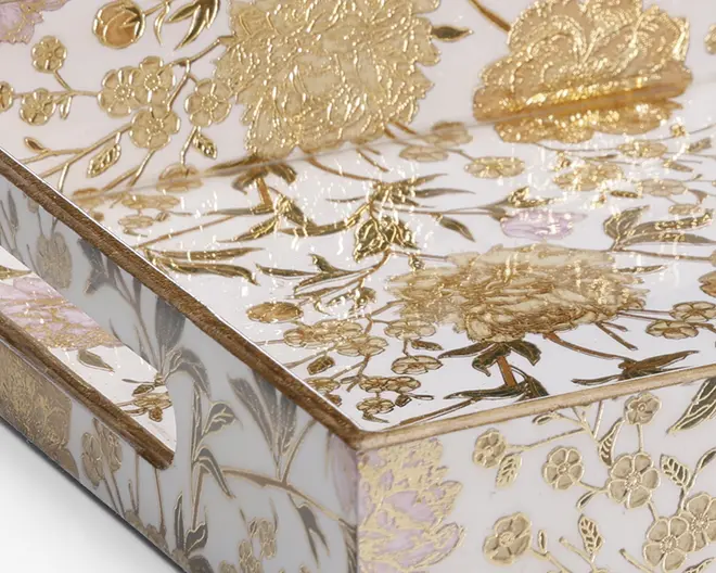 Nærbillede af et rektangulært objekt med en blank hvid overflade, dekoreret med detaljerede gyldne blomster- og bladmønstre samt præget relief. Blomsterne og bladene er fint markeret og hævet, hvilket giver et eksklusivt og tekstureret udseende. Kanterne er pænt afsluttede, og der er et udskåret håndtag på siden for nem transport.