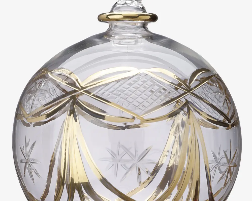 Klar glaskugle med indviklede guldfarvede båndmønstre og indgraverede stjernemotiver. Overfladen har både glatte og teksturerede områder, hvor gulddetaljer fremhæver de dekorative elementer. Toppen har en lille løkke til ophæng, også med gulddetalje.